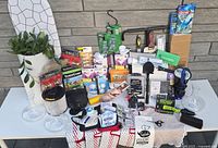 Wide view of all items in the lot on white table against brick wall including potted plant, light bulbs, tools, hardware boxes, decanters, and accessories.