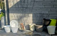 Photo showing an assortment of white, black, gray, and green plastic outdoor planting pots, some with built-in drainage and wheels, against a brick wall with wrought iron trellises in the background.