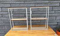 Two identical 3-tier wall shelves side by side on a wooden table with a gray brick wall background showing the front view of the shelves.
