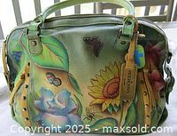 Front and side view of the green leather handbag featuring colorful hand-painted sunflowers, irises, butterflies, and bees, including the Anuschka signature tag and metal stud accents.