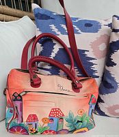 Full front view of the red leather satchel purse showing colorful hand-painted village houses and flowers, red handles and shoulder strap.