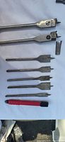 Several metal spade bits in various lengths and sizes laid flat showing sharp edges and measurement markings along with a red plastic tool.
