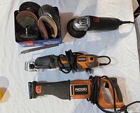 Overview of two orange and black Ridgid reciprocating saws, an electric angle grinder, and a tin of assorted grinding and cutting discs showing various disc types.