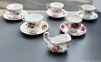 Six floral patterned Royal Albert teacups each with a matching saucer plus a cream pitcher on display outdoors.