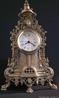 Front view of ornate brass mantel clock showing intricate latticework, decorative details, and four lion paw feet.