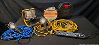 Wide shot of all items: 2 handheld work lights with protective cages and long cords (blue and yellow), yellow dual-head halogen floodlight, yellow retractable extension cord reel, spool of outdoor wire, heavy yellow extension cord, black surge protector power bar.