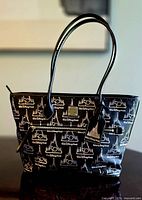 Front full view showing black coated cotton tote with white Walt Disney World 50th Anniversary and castle print, leather handles, front zip pocket with gold-tone zipper and attached holographic castle tag.