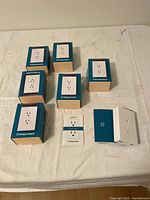 Seven boxed ThingCharger wall outlet chargers arranged on a flat surface with one unit removed from box showing front face with two power outlets.