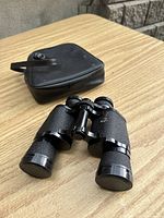 Full view of black Aerolite binoculars with closed lens caps and black carrying case on wood surface.