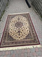 Full view of the Ankara woven tapestry rug laid flat outdoors on a stone textured ground, showing the overall decorative medallion floral pattern with beige, burgundy, and brown colors, and fringe on one side.