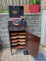 Open phonograph with wood cabinet showing multiple record storage shelves inside the door and an open drawer beneath the turntable