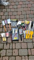 Overview of the entire assortment of painting tools laid on a brick surface showing paintbrushes, putty knives, rollers, utility knife, and trowel.