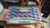 Top-down view of BaByliss hair curling set with colorful rollers under clear plastic cover, showing arrangement and size variety of rollers.