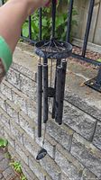 Black metal wind chime held by hand outdoors against stone wall and metal railing, showing hanging chimes and wind catcher.