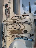 Top-down photo showing majority of the 20 assorted costume necklaces laid out on a countertop near a sink. Various bead styles, pendants, and charm necklaces are visible.