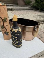 Photo showing all three items on a surface outdoors with clear view of woven bottle wrapped in black and yellow material, copper-tone oval beverage tub with rope handles, and tall terracotta vase with grapevine and swirl texture details.
