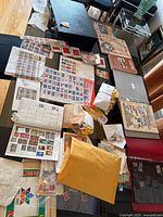 Photo of a table covered with multiple stamp albums, binders, envelopes, and loose stamp sheets spread out displaying stamps from various countries and eras including commemorative sheets and vintage covers.