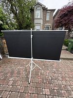 Projector screen with black backing fully extended on white tripod stand outdoors on brick.