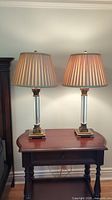 Pair of crystal lamps on wooden table with beige pleated lampshades, crystal octagonal columns, bronze ornate bases, shown plugged in and lit.