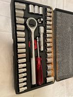 Open view of the portable socket set showing chrome sockets, ratchet, extension bar, and red-handled screwdriver in a black plastic case with foam interior showing rust marks inside the lid.