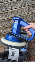 Blue Simoniz random orbit polisher with black and white pad attached, being held by a hand, with coiled power cord.