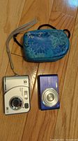 Two digital cameras (Sony Cybershot blue and HP Photosmart silver) placed on wooden floor along with blue carrying case
