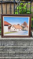 Full view of the oil painting showing the village street scene with houses, road, trees, and sky, set in a dark wood frame.