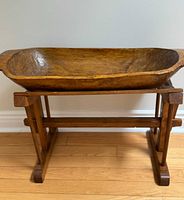Front view of large hand-carved wooden dough bowl on custom wood stand against a white wall on hardwood floor.