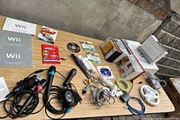 Wide shot of Nintendo Wii console, game cases stacked, controllers including guitars, microphones, and cables on a table.