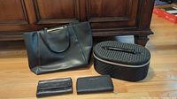 Set of a large black Kate Spade purse, two wallets (one Kate Spade and one Michael Kors), and a gray patterned cosmetic bag arranged in front of wood paneling.