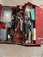 Open metal toolbox showing multiple tiers filled with assorted hand tools including wrenches, pliers, a small plastic organizer box, and other small hardware items.