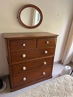 Solid wood chest of drawers in natural finish with five drawers and white knobs, shown standing with round mirror mounted behind.