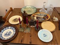 Overview photo of the lot on wooden table including decorative plates, brass candle holders with red candles, glass decanters, brown bottle, cat figurine, and large decorative bowl.