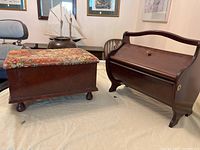 Side-by-side display of two wooden foot stools on a light-colored cloth. One has a floral fabric cushioned top; the other has a dark wooden curved handle and closed front compartment.