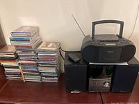 Photo showing the Sony stereo system with two speakers, a remote control, a Sony portable boombox on top of one speaker, and multiple stacks of assorted CDs on the left side.