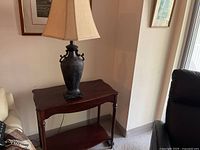 Side wooden table with dark finish and decorative metal lamp with beige shade on top, against corner of room.
