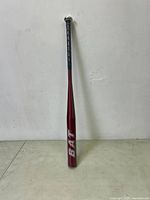 Full length view of the red aluminium baseball bat with black grip on handle leaning against a wall and floor.