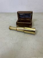 Solid brass handheld telescope placed next to open wooden storage box showing bubble wrap interior.