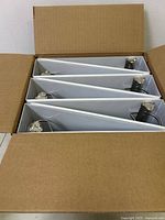 Top view of six white binders arranged inside a cardboard box showing the metal A angle D rings.
