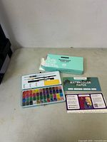 Full view of opened watercolor paint palette with 48 colors, two paint brushes, yellow sponge, Artistro watercolor paper pad, and the outer mint green box.