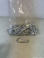 Clear plastic bag filled with silver metal carabiner clips and one clip placed outside the bag for display
