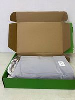 Green cardboard box opened showing light grey fitted sheet folded inside with a white cable and grounding device visible.
