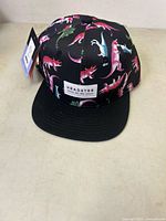 Front view of a black snapback hat with colorful dinosaur print and Headster logo patch on front.