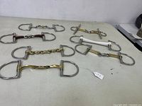 Seven assorted horse bits laid on a flat white surface, showing various mouthpiece styles including chain link, twisted wire, metal bars, and a white rubber piece.