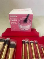 Red fabric pouch rolled out with 8 professional gold-ferruled makeup brushes placed inside, various sizes and shapes, synthetic bristles. Pink and white box labeled makeup and paint brush cleaner behind the pouch.
