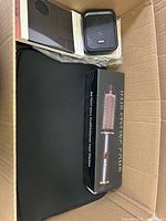 Open cardboard box showing a boxed hair styling comb, a small black portable speaker, a black tablet or laptop sleeve or case, and some other packaged items.