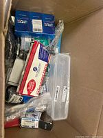 Box filled with various new and unopened items including Betty Crocker parchment paper, blue boxed paper products, scissors, and a clear plastic container.