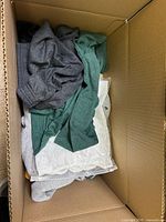 Top view inside cardboard box showing folded sweaters and tops in mainly grey, green, and white colors.