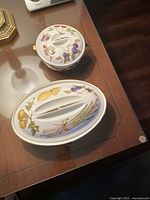 Two ceramic covered dishes on a glass table, showing oval and round shapes with floral and fruit patterns.
