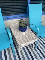 Photo shows a white wicker rectangular side table positioned between two bright blue outdoor chairs on a striped outdoor rug. The table has woven wicker legs and a flat woven surface. A dark blue planter with purple flowers is placed on the table, illustrating outdoor use and scale.